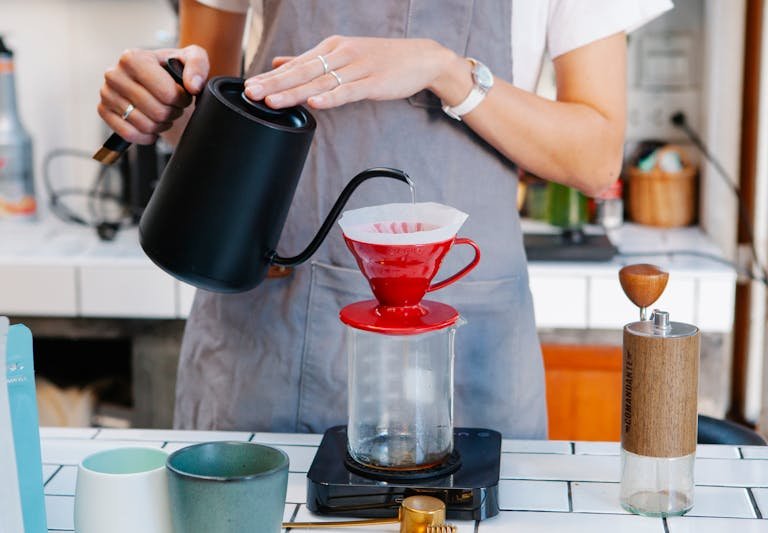 Crop unrecognizable female in gray apron brewing coffee with filter dripper in light kitchen in daytime